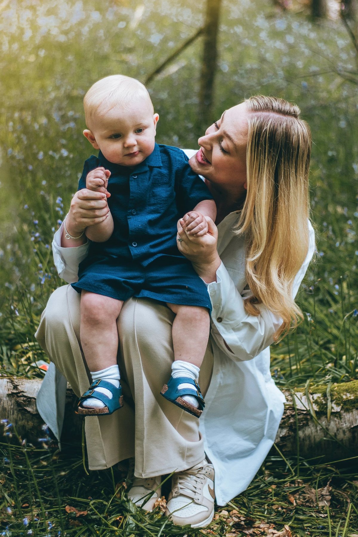 A baby in a white outfit sitting in a wicker basket set down among bluebells, laughing upward