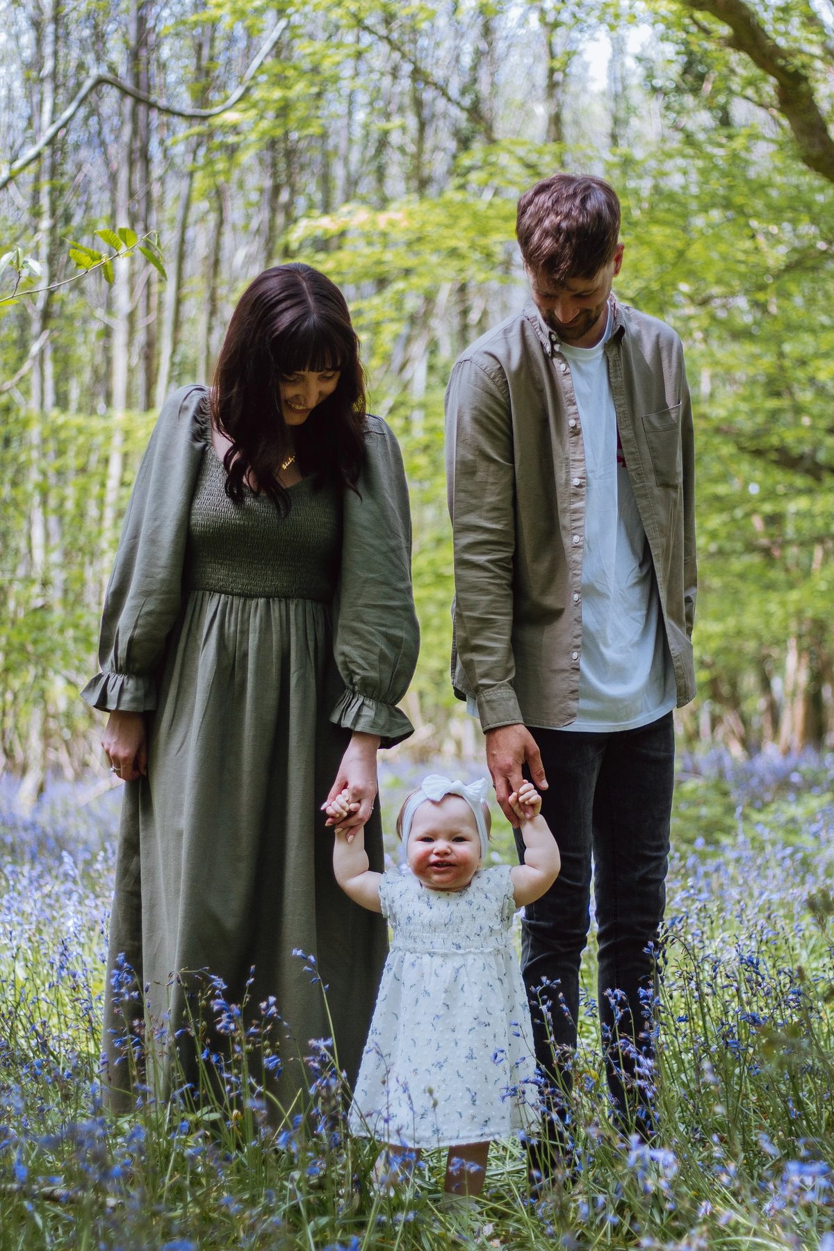 Two parents standing in a carpet of bluebells holding a toddler in a white dress between them