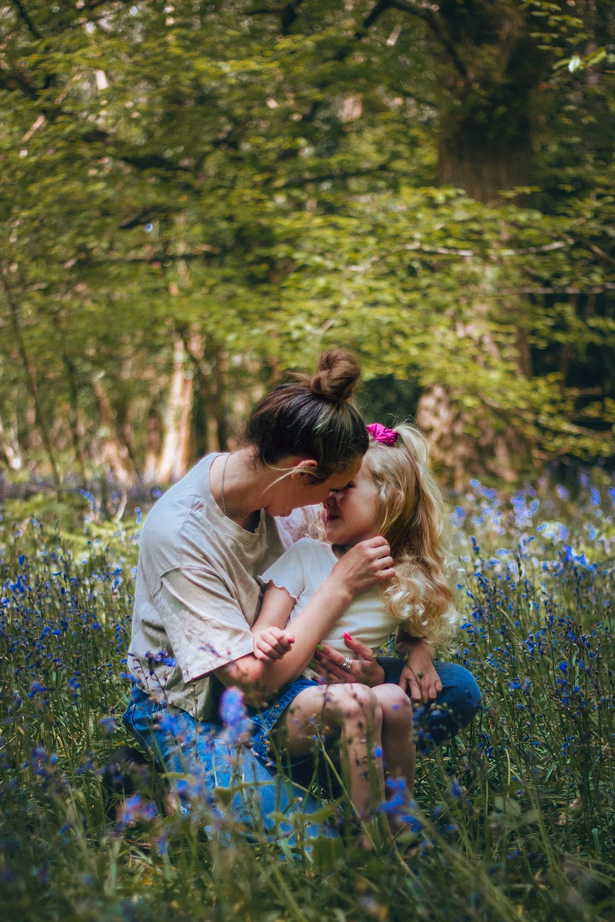 A mother crouched among bluebells kissing her small daughter, dappled woodland light behind them