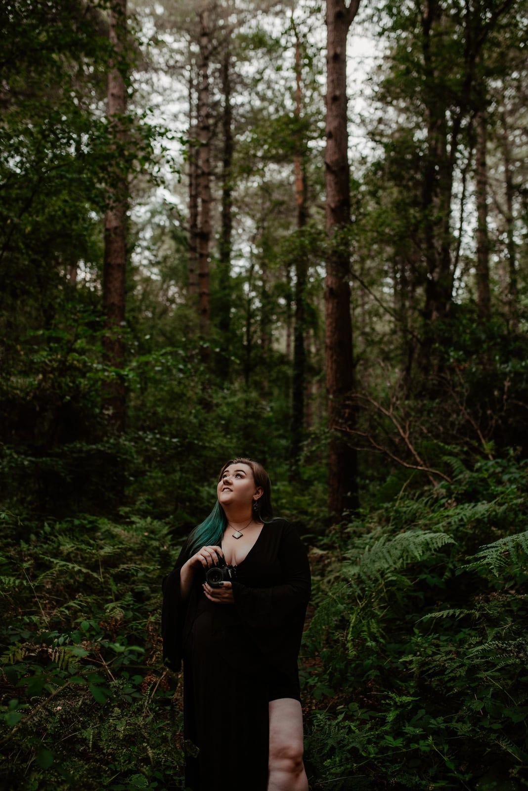Heather standing among ferns in a pine wood in a long black dress, camera in her hands, looking up at the light through the trees