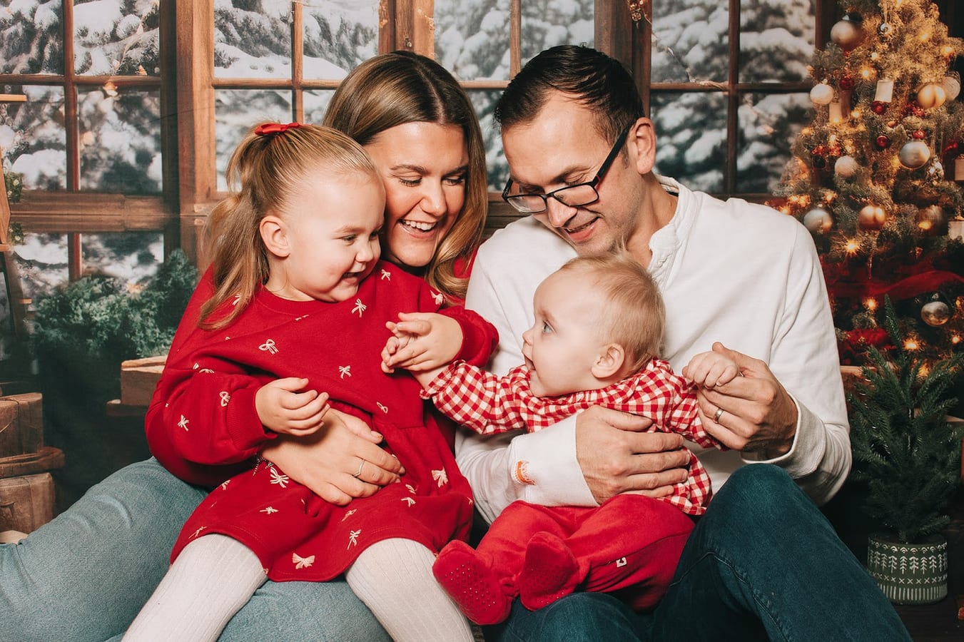 Two parents laughing with a toddler and a baby in front of a lit Christmas tree