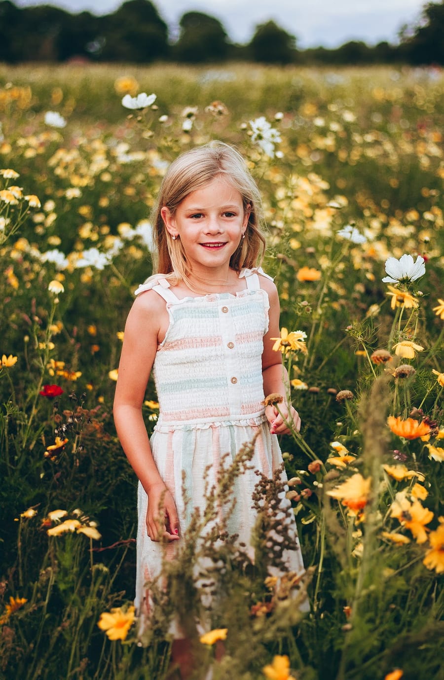 A young girl in a striped summer dress standing among yellow and white wildflowers at golden hour